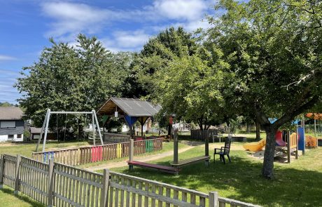 Bild sonniger Außenbereich Kita Höchstenbach mit Lattenzaun, Spielgeräten und Bäumen